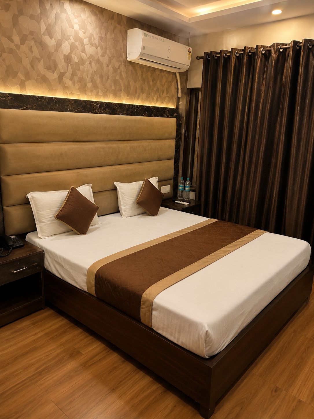 A hotel room with a large bed, a headboard, an air conditioning unit, a bedside table with a phone, and a nightstand.