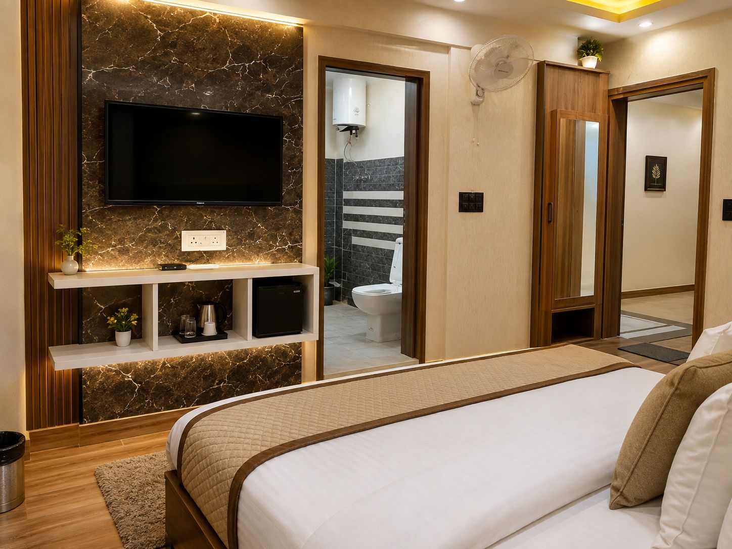 Un dormitorio moderno con una cama grande, un televisor montado, un estante con adornos y un baño visible a través de una puerta abierta.
