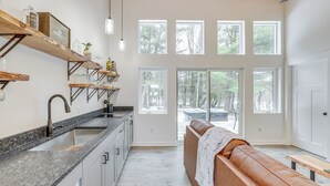 Une cuisine moderne avec un plan de travail en granit, un évier en acier inoxydable et des étagères en bois. Une grande fenêtre offre une vue sur les arbres, et un banc avec une couverture est installé à proximité.