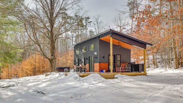Une cabane moderne dotée d’un porche abrité, entourée d’un paysage enneigé et d’arbres aux couleurs automnales.