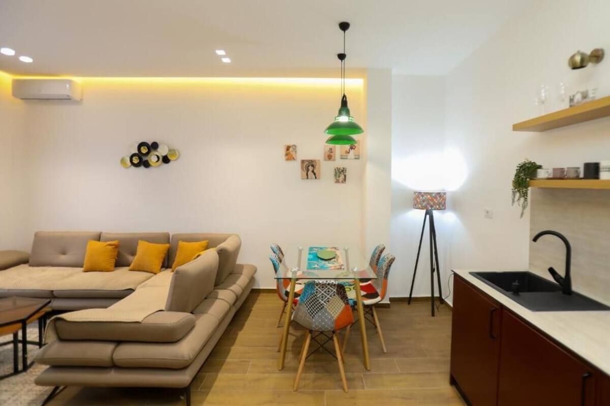 A modern living room with a sofa, a dining table with chairs, a kitchen area with a sink, and a pendant light.