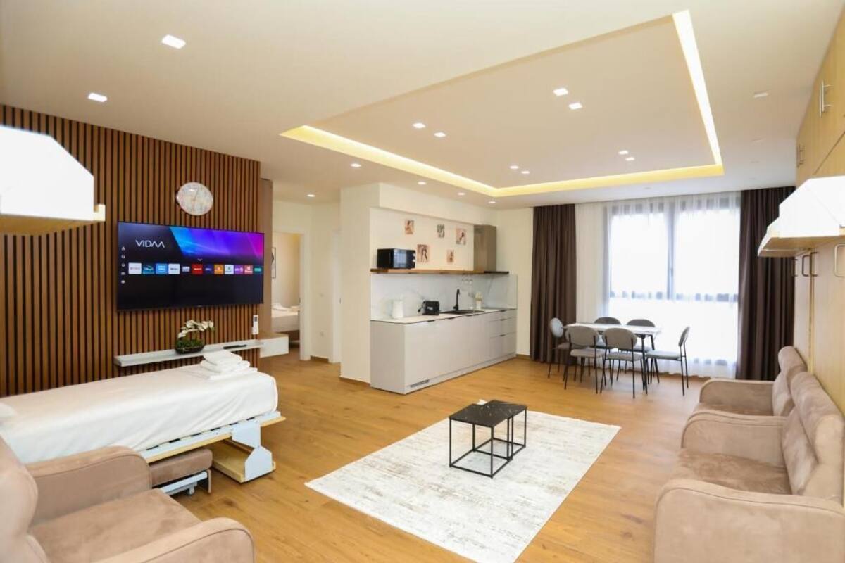 A modern hotel room with a bed, a TV, a kitchenette, a dining area, and a living space.
