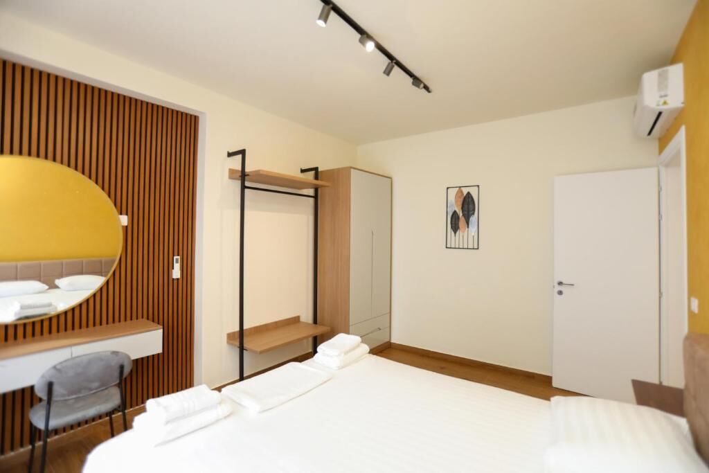 A hotel room with a bed, a desk with a chair, a wardrobe, a mirror, and an air conditioning unit.