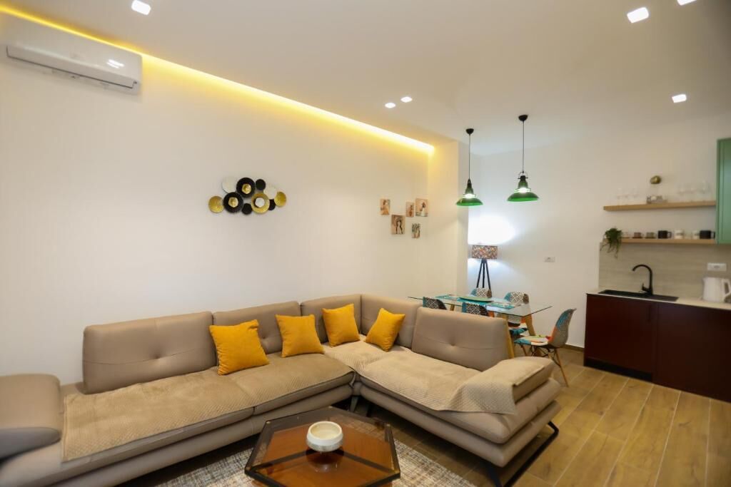 A modern living room with a sectional sofa, a coffee table, pendant lights, and a kitchen area in the background.