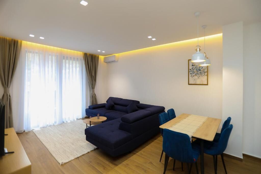 A modern living room with a sectional sofa, a dining table with chairs, a small side table, and a large window with curtains.
