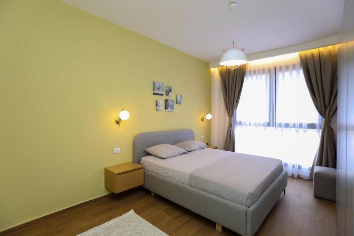 A bedroom with a bed, bedside tables, a nightstand, a lamp, a window with curtains, and wall art.