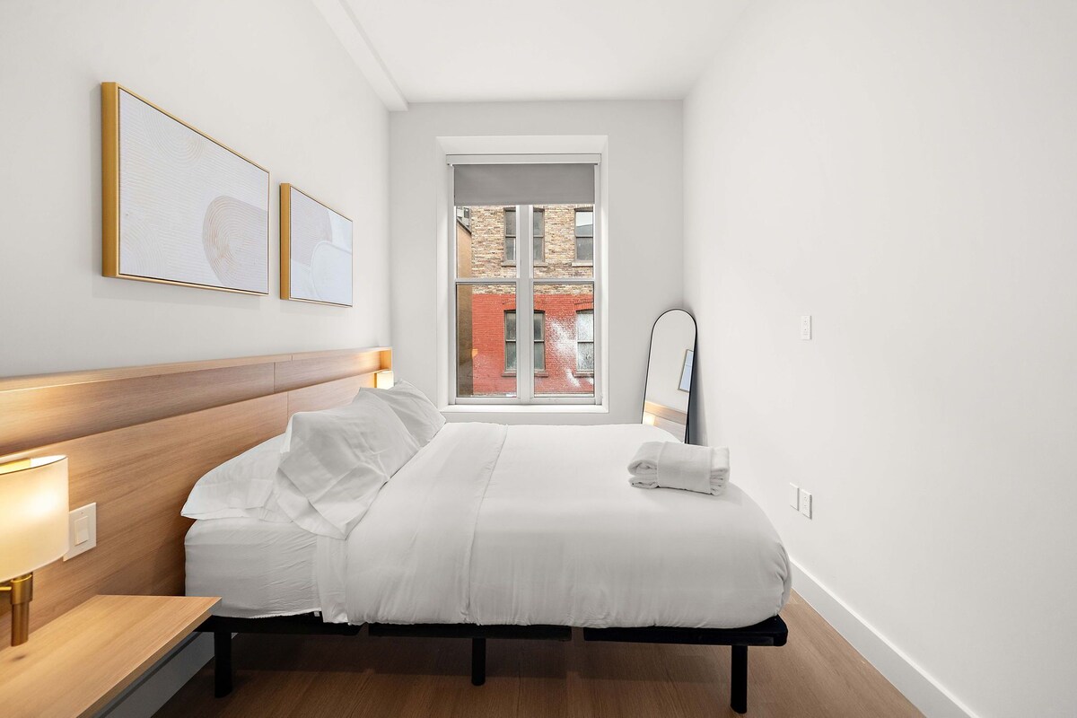 Ein Hotelzimmer mit Bett, Nachttisch, Lampe, Kunstwerk, Fenster und Spiegel.