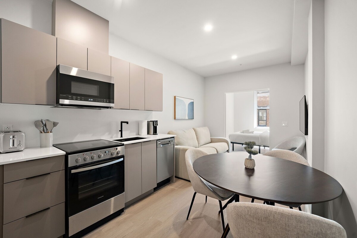 Eine moderne Küche mit Edelstahlgeräten, eine Essecke mit einem runden Tisch und ein Wohnbereich mit Sofa und Fernseher.