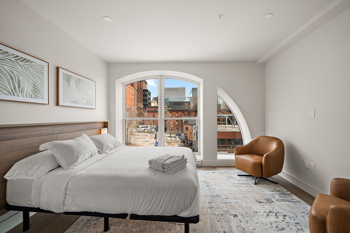 Ein modernes Schlafzimmer mit einem großen Bett, einem braunen Ledersessel und Blick auf eine Stadtkulisse durch ein großes Fenster.