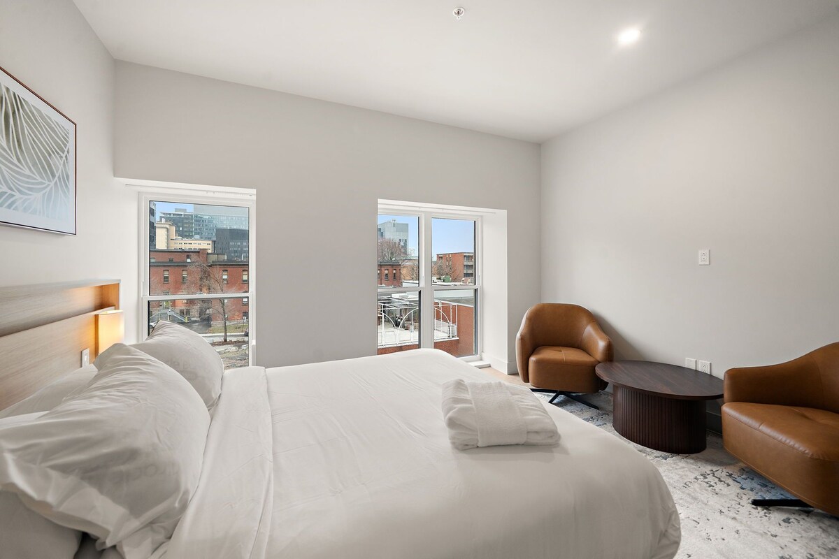 Ein Hotelzimmer mit einem großen Bett, zwei Sesseln, einem kleinen Tisch und Blick auf die Stadt durch die Fenster.