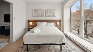A modern hotel room with a large bed, two bedside lamps, a flat-screen TV, and a large window offering a city view. - Nomade Saint Paul (Montréal)