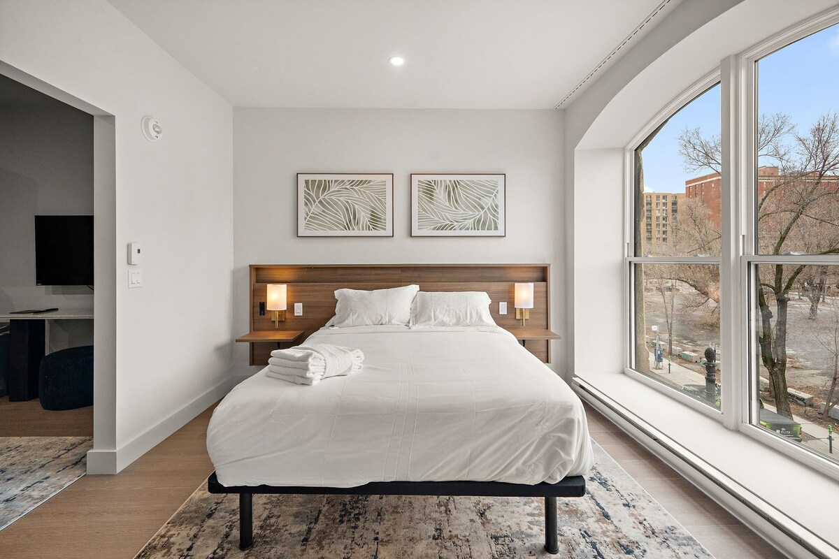 Ein modernes Hotelzimmer mit einem großen Bett, zwei Nachttischlampen, einem Flachbildfernseher und einem großen Fenster mit Blick auf die Stadt.