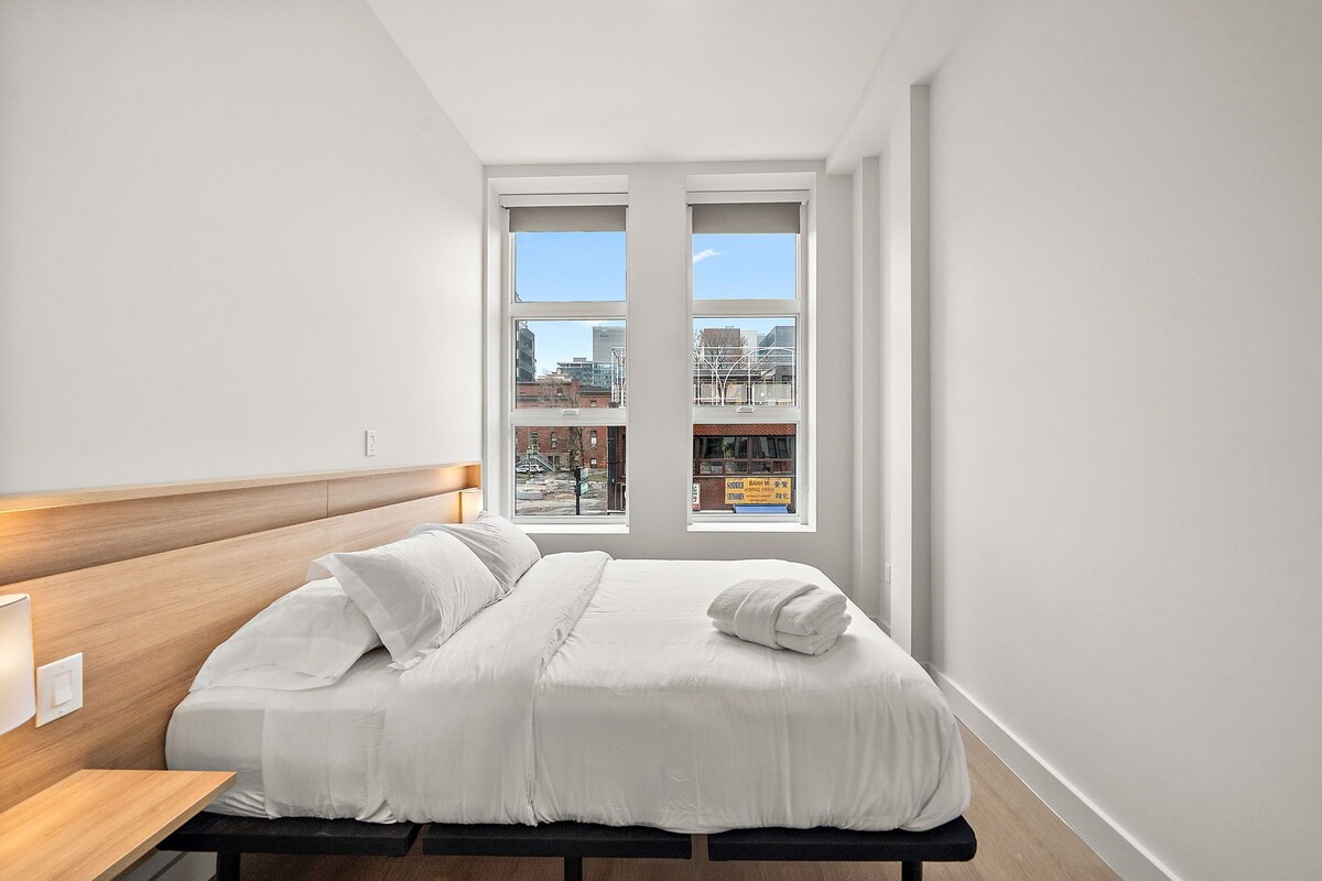 Ein modernes Hotelzimmer mit einem großen Bett, einem Holzkopfende, einem Nachttisch und Blick auf die Stadt durch große Fenster.