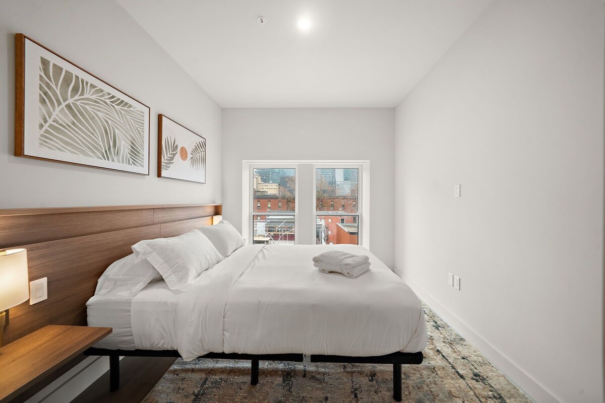 Ein Schlafzimmer mit Bett, Nachttisch, Lampe, Kunstwerk und Blick auf Gebäude durch ein Fenster.
