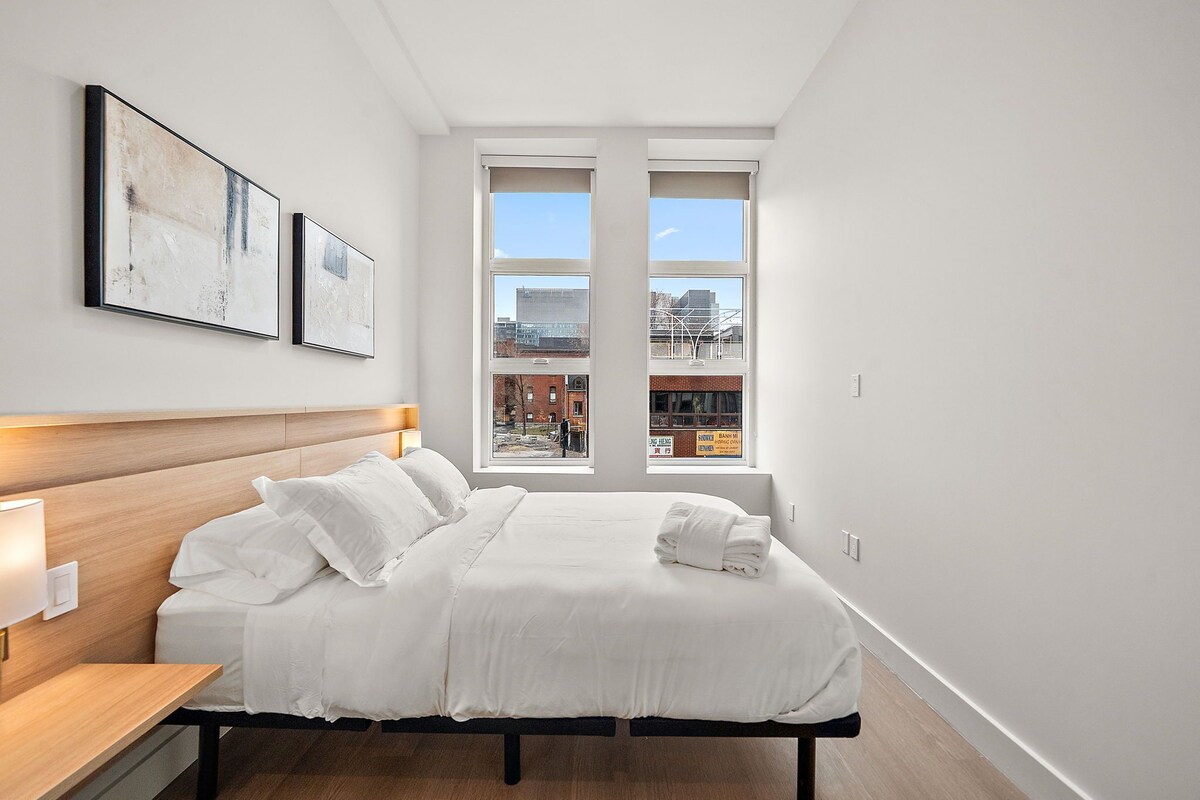 Ein Hotelzimmer mit einem großen Bett, einem Kopfteil, einem Nachttisch mit Lampe und einem Fenster mit Blick auf die Stadt.