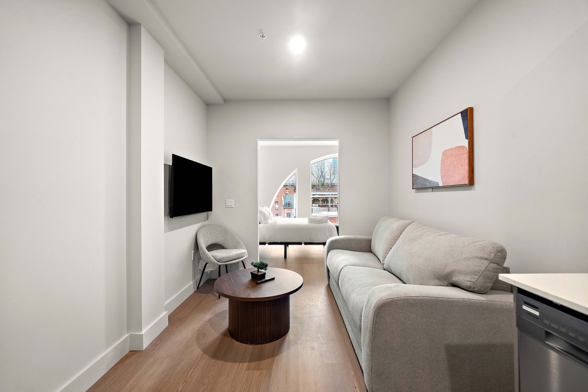 Un soggiorno moderno con divano, poltrona e tavolino, una TV a schermo piatto montata a parete e una finestra con vista sugli edifici.