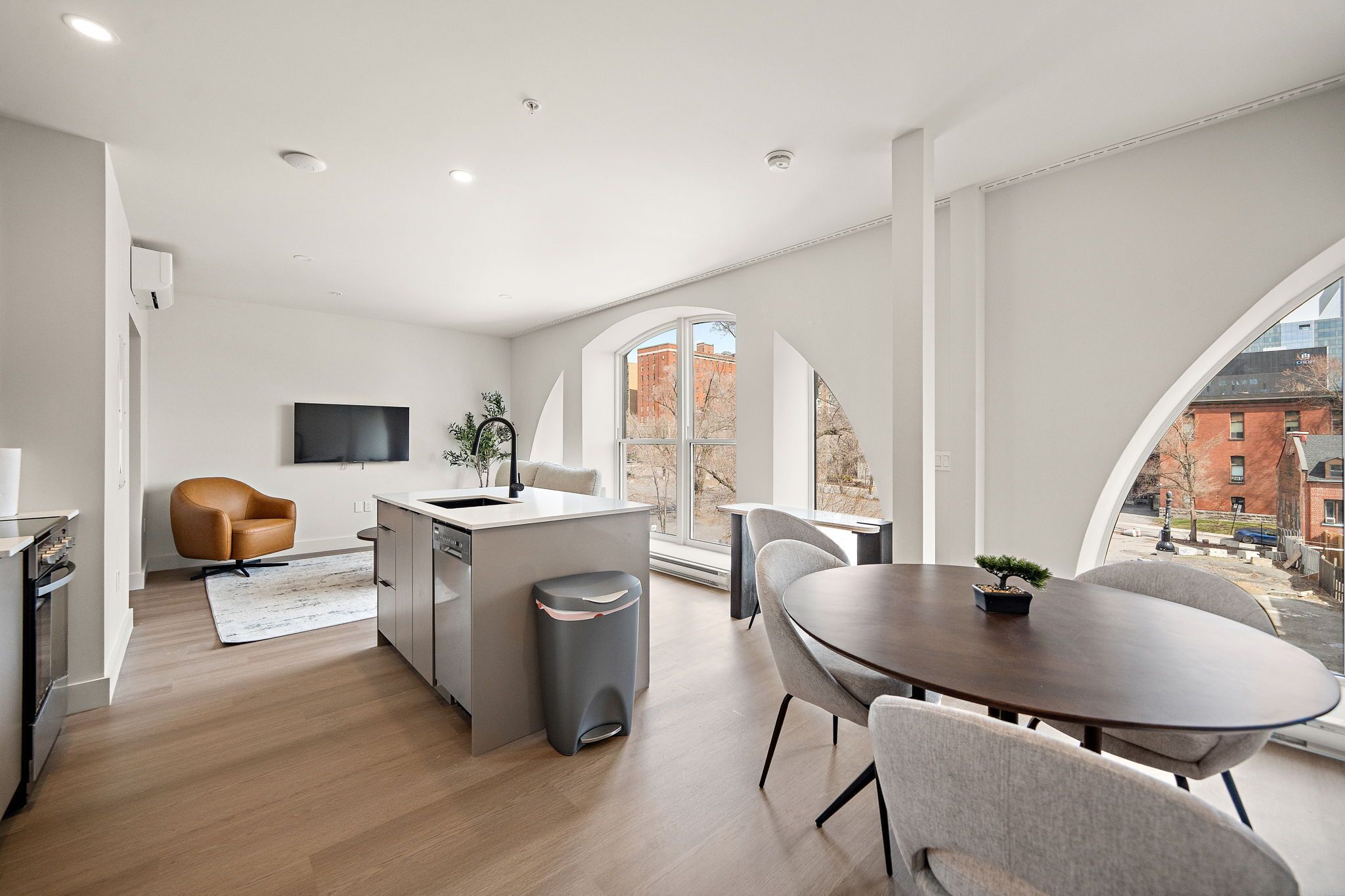 A modern kitchen with a central island, a dining area with a round table, and large windows offering a view of buildings.