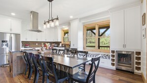 A modern kitchen with white cabinets, a central island with seating, stainless steel appliances, and a large window with a view of greenery. - 5BR Cabin on 3 Acres w Theater, Hot Tub +Fire Pit (Boone)