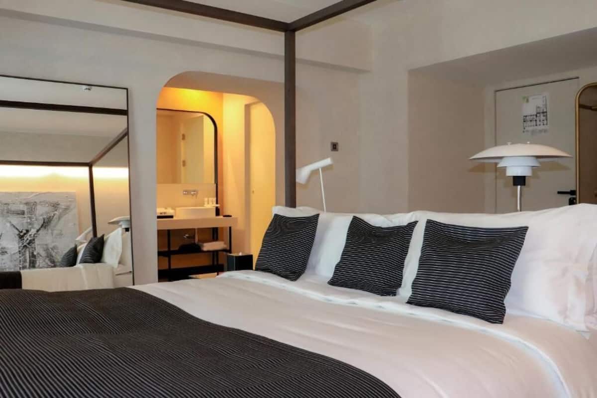 Ett hotellrum med en säng, ett skrivbord, en spegel, en lampa och ett badrum som syns genom en valvöppning.