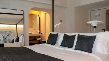 A hotel room with a bed, a desk, a mirror, a lamp, and a bathroom visible through an archway.