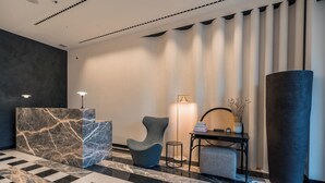 A lobby with a marble reception desk, a modern armchair, a small side table, a large cylindrical planter, and a checkered ottoman. - Monograph Freedom Square (Tbilisi)