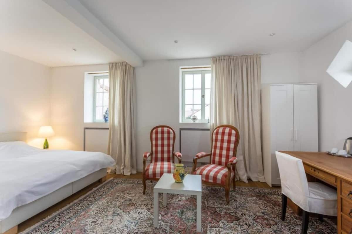 Ein Hotelzimmer mit einem Bett, zwei karierten Sesseln, einem kleinen Tisch mit einer Karaffe, einem Schreibtisch mit Stuhl und einem Kleiderschrank.