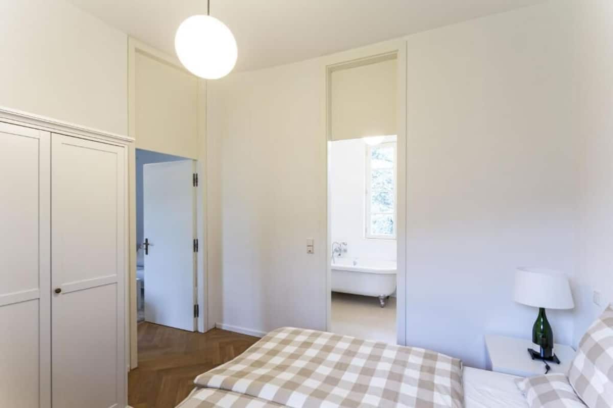 Ein Schlafzimmer mit karierter Bettdecke, einem Nachttisch mit Lampe, einem weißen Kleiderschrank und Blick auf ein Badezimmer mit Badewanne.