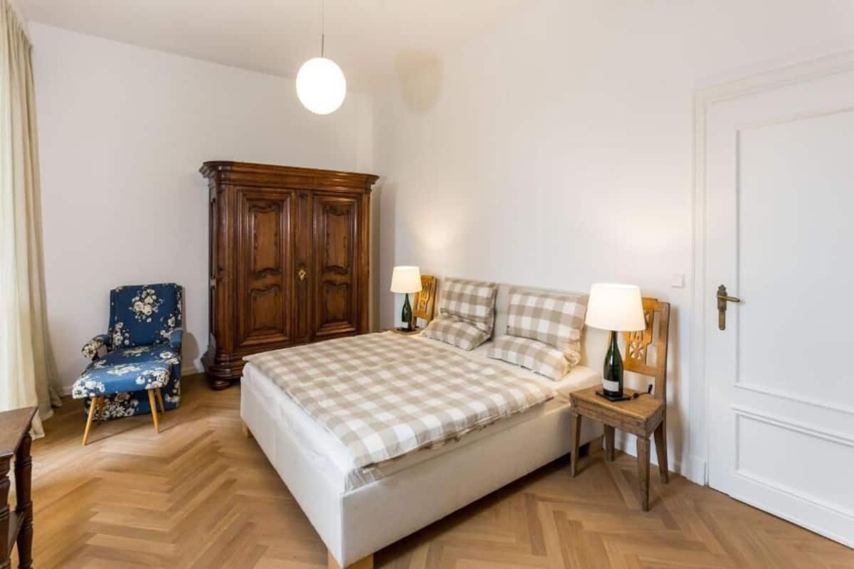 Ein Schlafzimmer mit einem Bett, zwei Nachttischen mit Lampen, einem Holzschrank, einem blumigen Sessel und einem Parkettboden mit Fischgrätmuster.