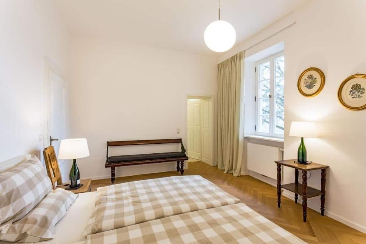 Ein Schlafzimmer mit einem Bett, einer Bank, einem Tisch und einem Fenster mit Vorhängen.