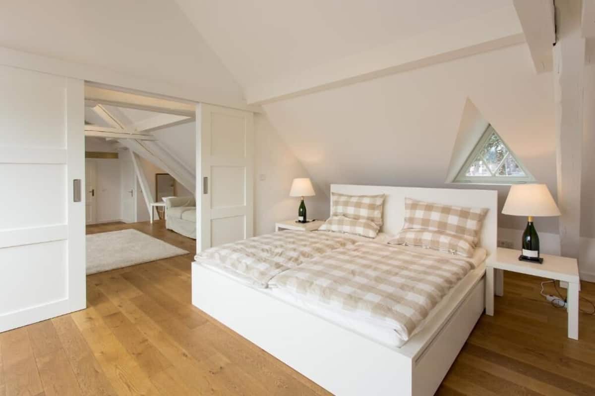 Ein Schlafzimmer mit einem großen Bett, zwei Nachttischen, einem Fenster und einer Treppe, die in einen anderen Raum führt.