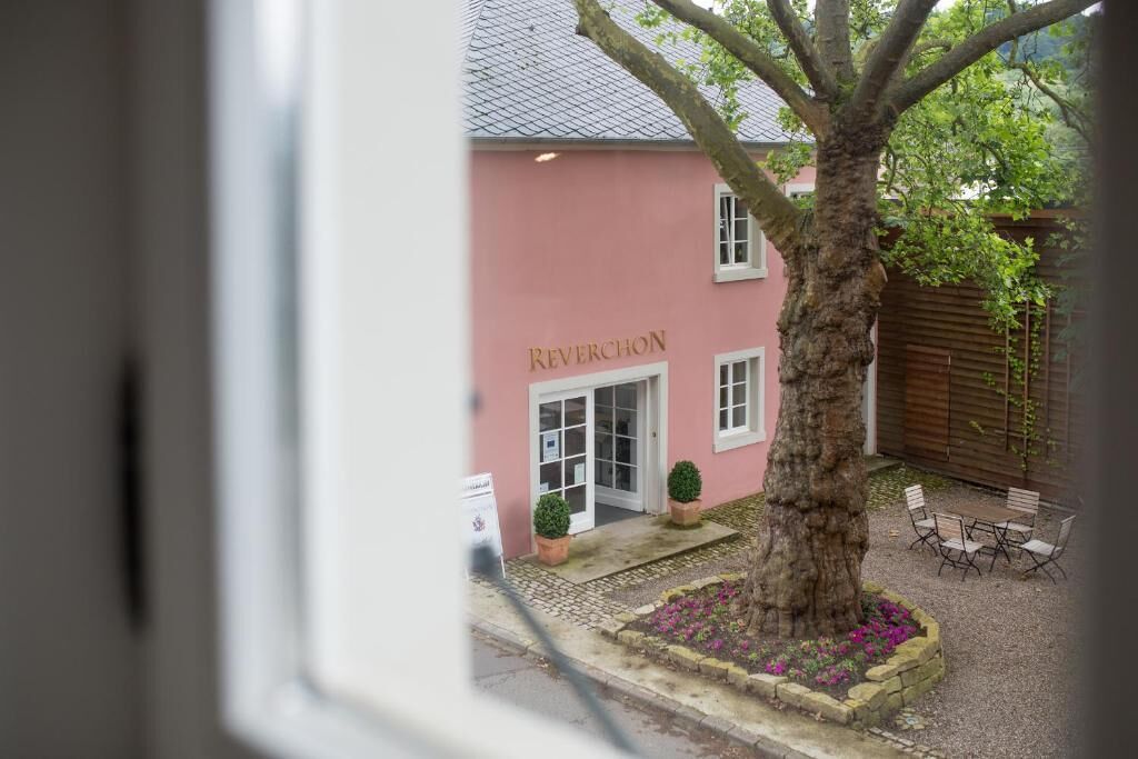 A building with a pink facade and the name 'REVERCHON' visible. There is a tree in the center, a patio with chairs, and a small garden area with flowers.
