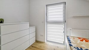 A room with a white dresser, a sliding door with blinds, a radiator, and a bed with a patterned bedspread. - Apartment 2 (Right) (Mössingen)