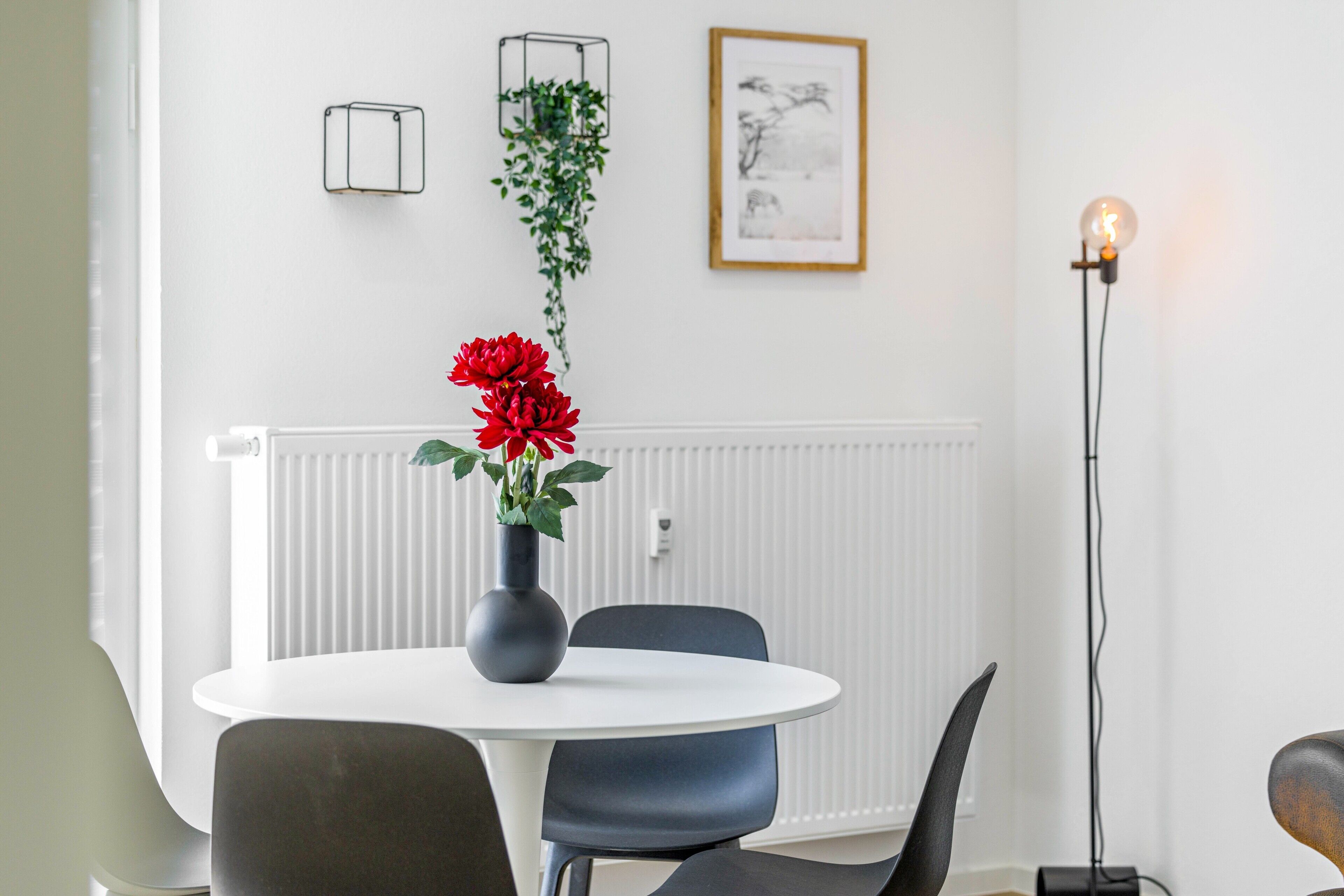 A dining area with a round table, chairs, a vase with flowers, a radiator, a framed picture, and a floor lamp.