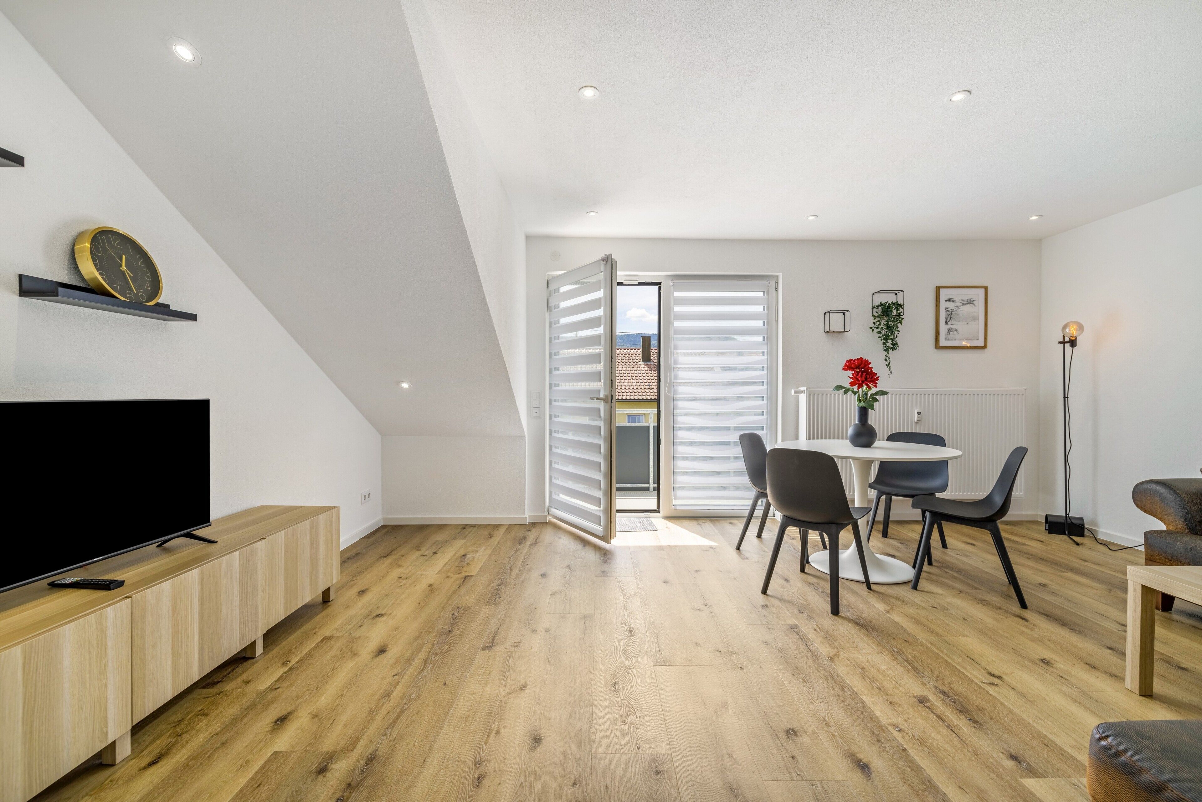 A modern living room with a flat-screen TV, wooden flooring, a dining area with a table and chairs, and a view through a door.