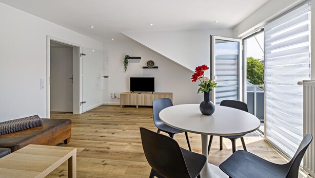 A modern living room with a wooden floor, a round table with chairs, a sofa, a TV, and a sliding glass door leading to a balcony. - Apartment 2 (Right) (Mössingen)