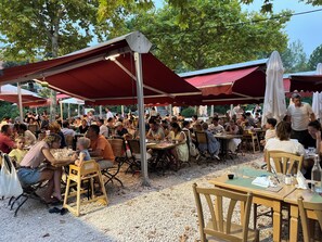 An outdoor dining area with tables and chairs, covered by red canopies, surrounded by trees. - House le Rouret (Grospierres)