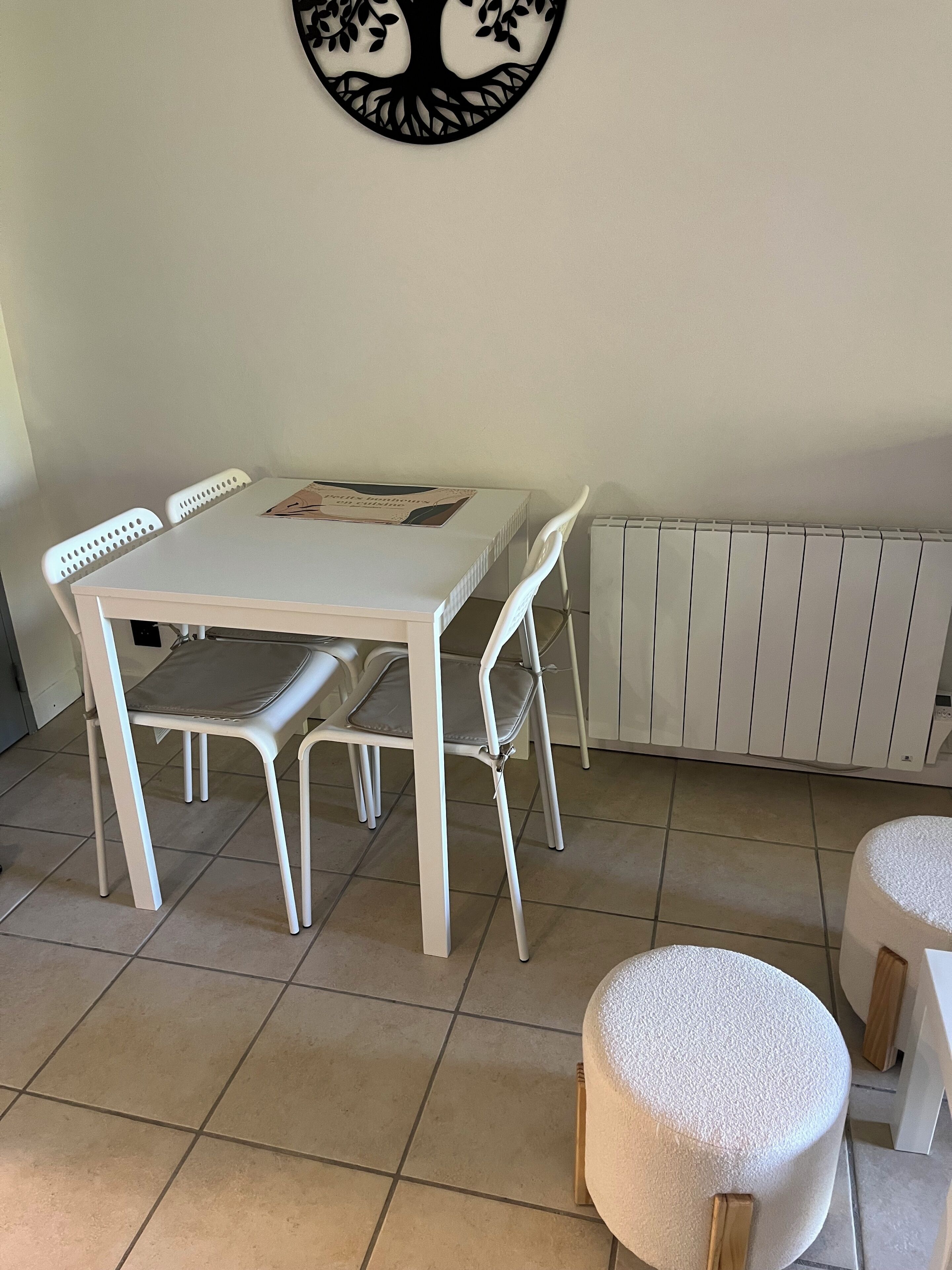 Een eethoek met een witte tafel, vier stoelen, een ronde decoratie aan de muur, een radiator en twee krukken.