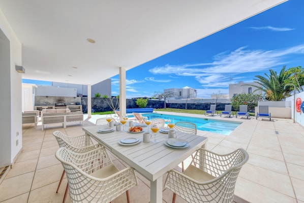 Un espace extérieur aménagé pour les repas, avec une piscine, des meubles blancs et un ciel d’un bleu limpide.