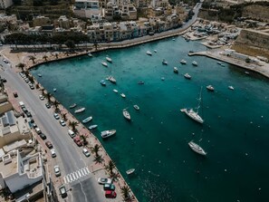Un port de plaisance situé sur la côte, avec des bateaux amarrés, une route qui le longe et des bâtiments en arrière-plan.
