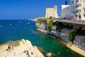 Uma vila costeira com um forte histórico, mar azul-turquesa e uma praia de pedras onde pessoas tomam sol.