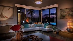 A modern living room with a sectional sofa, a red chair, a coffee table, a lamp, and large windows with a view of the evening sky. - Villa El Erizo • Pool • LVH (Nazaret)
