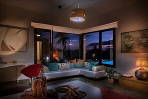 A modern living room with a sectional sofa, a red chair, a coffee table, a lamp, and large windows with a view of the evening sky. - Villa El Erizo • Pool • LVH (Nazaret)