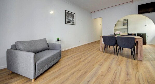 A modern living room with a grey sofa, a dining table with four chairs, a small side table with a plant, and a framed picture on the wall. - Modern St Paul`s Bay Apartment w/ Pool | Sleeps 4 (San Pawl il-Baħar)