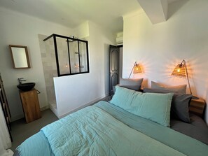 Quarto com cama, luminárias de cabeceira, janela e espelho, além de banheiro com chuveiro.