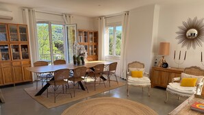 Ein Essbereich mit rundem Tisch, Korbstühlen, einer Speisekammer und Blick auf Grün durch große Fenster.