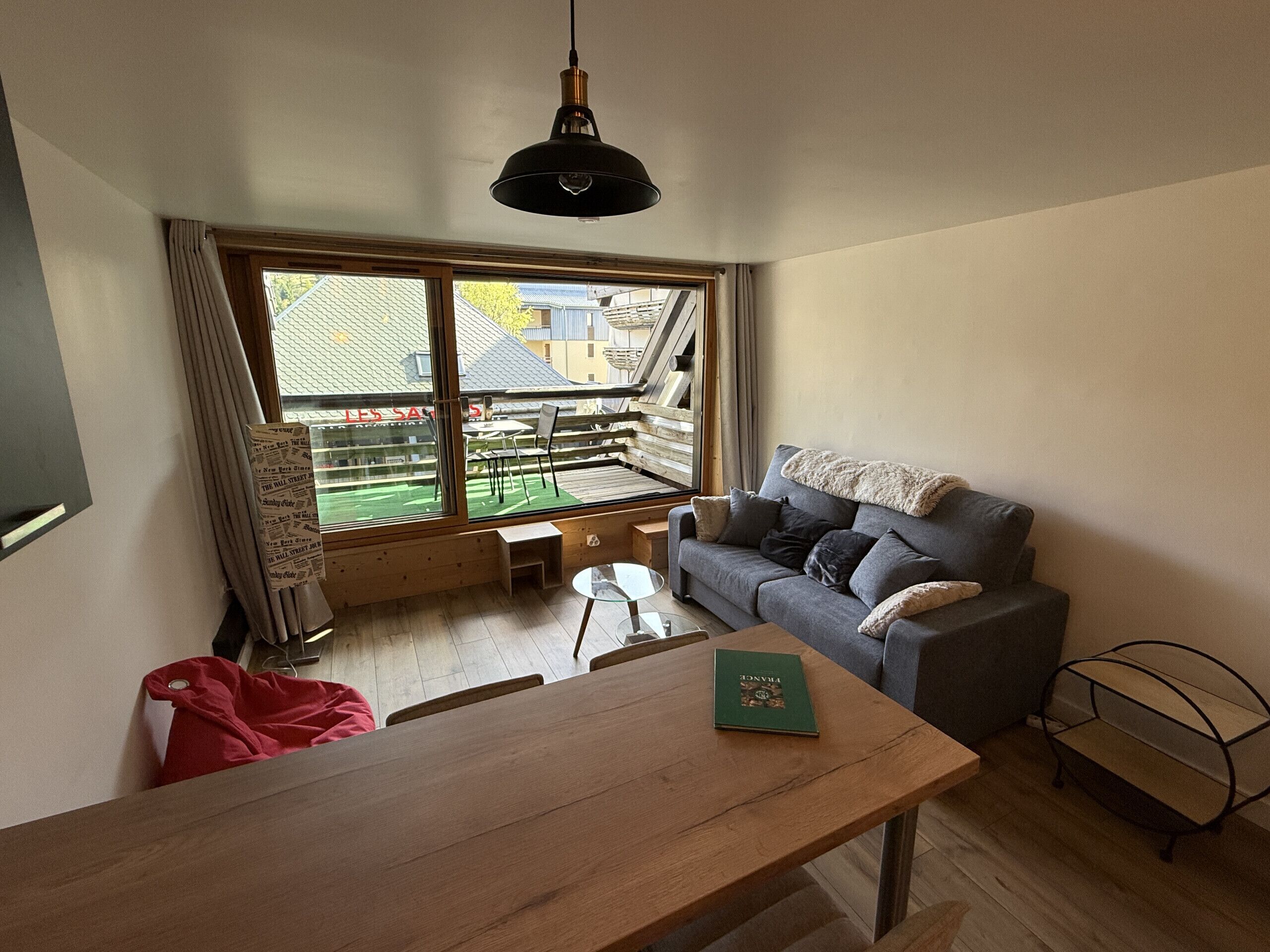 A room with a wooden table, a grey sofa, a window with a view of buildings, and a hanging light fixture.