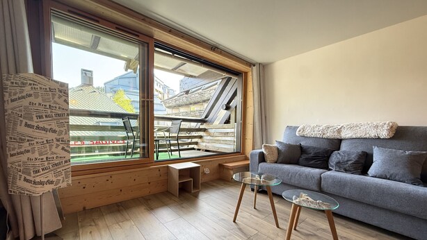 Een moderne woonkamer met een groot raam, een grijze bank, een glazen salontafel en een houten vloer.