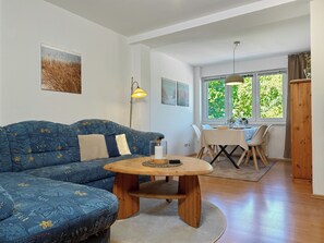 Un salon comprenant un canapé, une table ronde en bois, une table à manger avec des chaises, une lampe et une fenêtre donnant sur un paysage verdoyant.
