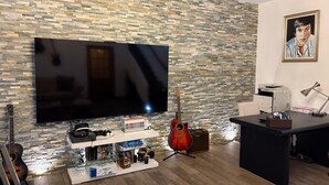 Ein Zimmer mit Steinwand, einem Flachbildfernseher, einer Gitarre, einem Schreibtisch mit Drucker und einem Bücherregal mit verschiedenen Gegenständen.