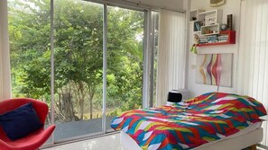 Un dormitorio con un ventanal grande, una cama con colcha de colores, una silla roja y una estantería con libros y objetos decorativos.